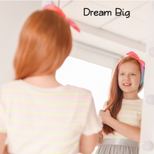 Load image into Gallery viewer, A young girl with long red hair and a colourful headband smiles into a mirror, wearing a striped top. The text "Dream Big" is in the corner, conveying optimism.