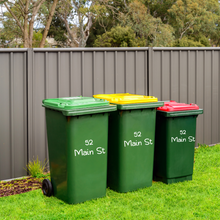 Load image into Gallery viewer, Three green garbage bins with green, yellow, and red lids are placed on a lawn in front of a grey metal fence. Each bin is labelled "52 Main St."