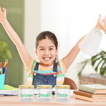 Load image into Gallery viewer, A young girl with raised arms sits at a table with three labelled jars: "Splurge", "Donate", and "Smile". A desk lamp and office supplies are visible in the background.