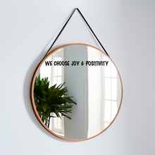 Load image into Gallery viewer, Round mirror with wooden frame hanging on a wall, reflecting a plant and windows. The decal text on mirror reads "We Choose Joy & Positivity.” Bright and uplifting.