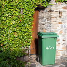 Load image into Gallery viewer, Green wheeled bin labelled "4/20" stands on stone path near a wooden door, surrounded by lush green ivy and rustic stone wall, conveying a serene garden atmosphere.