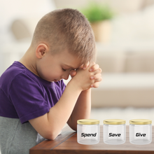 Load image into Gallery viewer, A boy in prayer position with three jars labelled "Spend," "Save," and "Give on a wooden table.