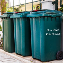 Load image into Gallery viewer, Three green garbage bins line a side street against a metal fence. One bin has the words "Slow Down Kids Around" written on it, conveying caution.