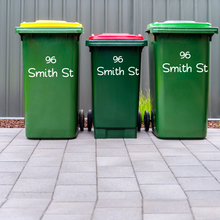 Load image into Gallery viewer, Three green garbage bins with coloured lids (yellow, red, and green) stand on a grey stone pavement against a metal fence, labeled "96 Smith St."