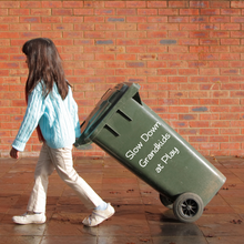 Load image into Gallery viewer, A girl in a blue sweater pulls a green trash bin with "Slow Down Grandkids at Play" on it. She walks on a tile path against a brick wall backdrop.