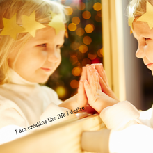 Load image into Gallery viewer, A young child with a yellow star crown gazes into a mirror, touching it gently. Warm, festive lights glow in the background. Text: "I am creating the life I desire."