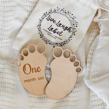 Load image into Gallery viewer, Wooden milestone markers shaped like baby feet with "One month old" and "Two month old" inscriptions next to a drawstring bag labeled "Live Laugh Label."