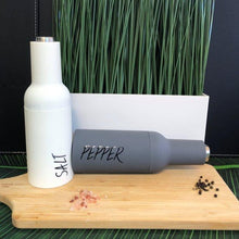 Load image into Gallery viewer, White salt and gray pepper grinders labelled "SALT" and "PEPPER" on a wooden board with pink Himalayan salt and peppercorns, set against tall green grass.