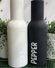 Load image into Gallery viewer, Two modern, cylindrical salt and pepper shakers labelled "SALT" and "PEPPER." One is white, the other black, with greenery in the background.