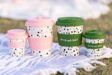 Load image into Gallery viewer, Four reusable coffee cups on a picnic blanket, featuring pink and green lids with polka dot patterns and a calm, outdoor setting. The branding "Live Laugh Label" on each cup.