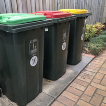 Load image into Gallery viewer, Three green wheelie bins with lids in red, yellow, and green stand on a paved area by a wooden fence and grass, marked with "Brisbane City" and a "10" decal on the front representing the unit number.