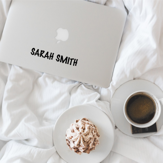 Picture of a laptop with a decal of the name Sarah Smith on a bed with a coffee and treat to eat